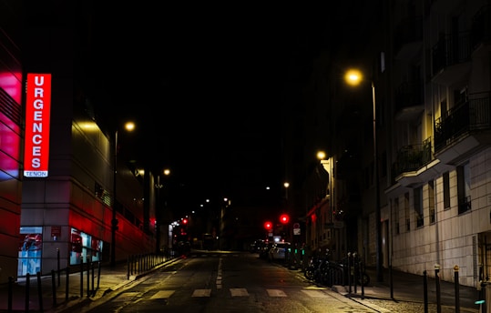 A nighttime urban street scene featuring an illuminated emergency sign with the word 'Urgences' in bright red letters on a building. Streetlights cast a warm glow over the wet pavement and the surrounding buildings are mostly in shadow. The street is empty, adding a sense of quiet and stillness.