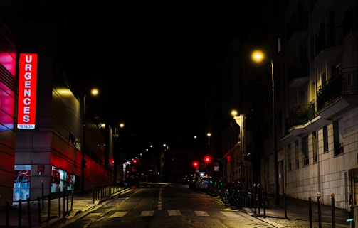A nighttime urban street scene featuring an illuminated emergency sign with the word 'Urgences' in bright red letters on a building. Streetlights cast a warm glow over the wet pavement and the surrounding buildings are mostly in shadow. The street is empty, adding a sense of quiet and stillness.