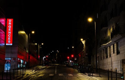 A nighttime urban street scene featuring an illuminated emergency sign with the word 'Urgences' in bright red letters on a building. Streetlights cast a warm glow over the wet pavement and the surrounding buildings are mostly in shadow. The street is empty, adding a sense of quiet and stillness.
