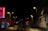 A nighttime urban street scene featuring an illuminated emergency sign with the word 'Urgences' in bright red letters on a building. Streetlights cast a warm glow over the wet pavement and the surrounding buildings are mostly in shadow. The street is empty, adding a sense of quiet and stillness.