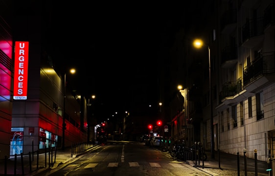 A nighttime urban street scene featuring an illuminated emergency sign with the word 'Urgences' in bright red letters on a building. Streetlights cast a warm glow over the wet pavement and the surrounding buildings are mostly in shadow. The street is empty, adding a sense of quiet and stillness.