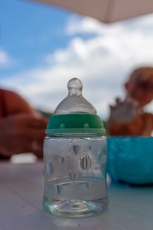 A baby bottle with a green cap featuring heart-shaped decorations is placed on a white surface. The background is blurred, showing a person holding an object and an umbrella against a partly cloudy sky.