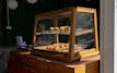 A wooden display case with glass panels containing various pastries, including donuts and other baked goods, is situated on a wooden counter. Behind the display, a person is visible, with a table containing a few items, potentially for business transactions.