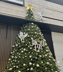 A large, decorated Christmas tree is adorned with white and gold ornaments. White angel figurines with wings are strategically placed around the tree. The word 'JINGLE BAY' is prominently displayed in white letters on the tree. A golden star tops the tree, and the background features modern building elements.