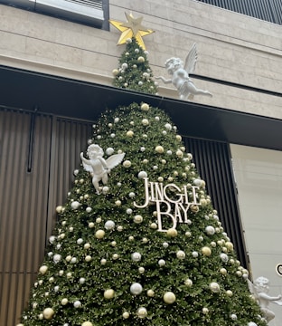 A large, decorated Christmas tree is adorned with white and gold ornaments. White angel figurines with wings are strategically placed around the tree. The word 'JINGLE BAY' is prominently displayed in white letters on the tree. A golden star tops the tree, and the background features modern building elements.