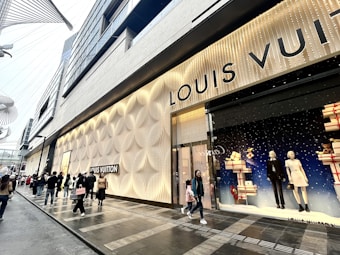 A storefront featuring the Louis Vuitton brand is adorned with elegant and textured wall designs. Large windows display mannequins dressed in fashionable outfits, surrounded by decorative gift boxes. A modern street scene with several people walking by, some carrying shopping bags, adds to the bustling city atmosphere.