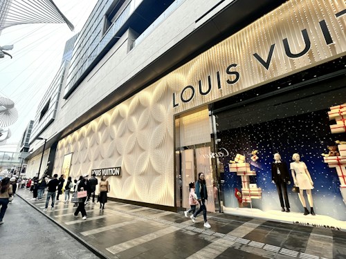 A storefront featuring the Louis Vuitton brand is adorned with elegant and textured wall designs. Large windows display mannequins dressed in fashionable outfits, surrounded by decorative gift boxes. A modern street scene with several people walking by, some carrying shopping bags, adds to the bustling city atmosphere.