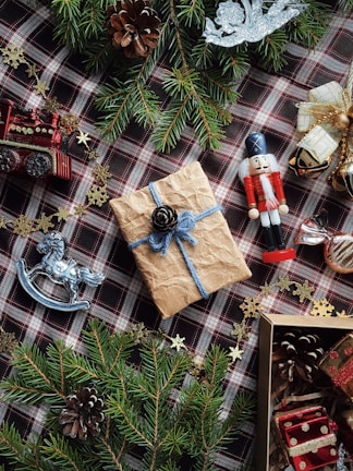 A festive arrangement featuring a wrapped gift with a pinecone on top, surrounded by evergreen branches, a toy nutcracker, pinecones, red gift boxes, and decorative items such as a silver rocking horse and a red train. Gold snowflake ornaments are scattered around a plaid fabric background, enhancing the holiday theme.