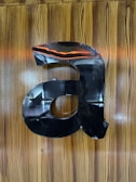 A large, black, metallic letter 'a' is mounted on a wooden surface with a glossy finish. The wood grain is vertically aligned, and there is a reflection of some lights and possibly a person in the shiny surface of the letter.