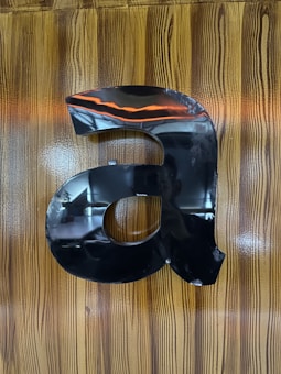 A large, black, metallic letter 'a' is mounted on a wooden surface with a glossy finish. The wood grain is vertically aligned, and there is a reflection of some lights and possibly a person in the shiny surface of the letter.