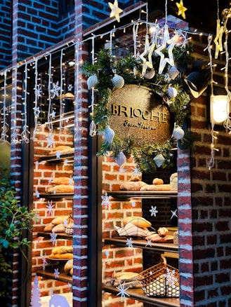 A festive bakery window display featuring a variety of breads on shelves against a brick wall. The store is decorated with hanging star lights and snowflake ornaments. A lush green wreath adorned with baubles surrounds a golden sign that reads 'BRIOCHE'. Warm lighting enhances the cozy atmosphere.