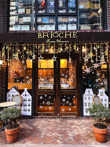 A cozy bakery storefront featuring wooden doors adorned with decorative stars and snowflakes. Above the doors, there are shelves filled with stacked boxes, and the shop name 'BRIOCHE From Heaven' is displayed in elegant gold lettering. Two potted plants flank the entrance, and a small table with a chair is placed to the left.