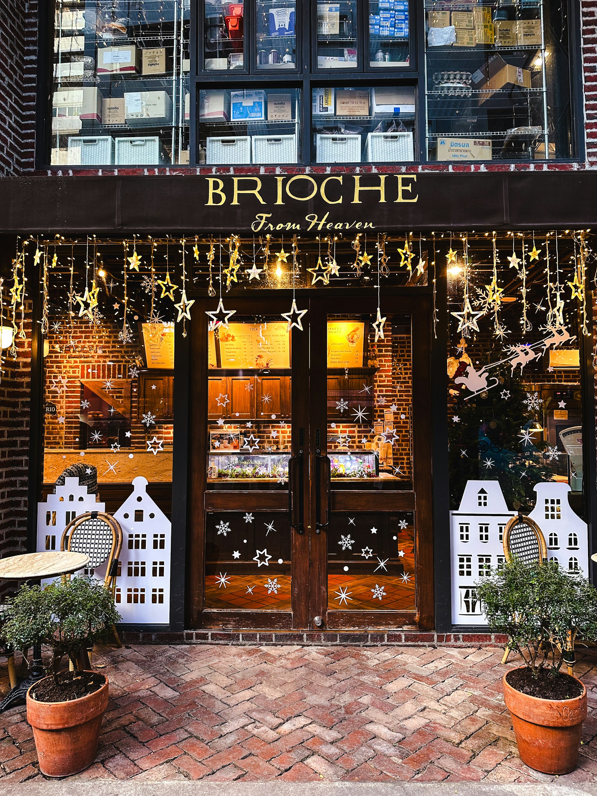 A cozy bakery storefront featuring wooden doors adorned with decorative stars and snowflakes. Above the doors, there are shelves filled with stacked boxes, and the shop name 'BRIOCHE From Heaven' is displayed in elegant gold lettering. Two potted plants flank the entrance, and a small table with a chair is placed to the left.