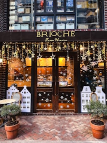 A cozy bakery storefront featuring wooden doors adorned with decorative stars and snowflakes. Above the doors, there are shelves filled with stacked boxes, and the shop name 'BRIOCHE From Heaven' is displayed in elegant gold lettering. Two potted plants flank the entrance, and a small table with a chair is placed to the left.