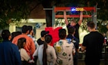 A man is performing a challenge where he hangs from a bar with a digital timer above him. A crowd gathers around, observing with interest and curiosity. The scene takes place outdoors, possibly at a fair or public event, with trees and lights in the background.