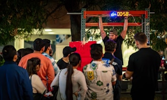 A man is performing a challenge where he hangs from a bar with a digital timer above him. A crowd gathers around, observing with interest and curiosity. The scene takes place outdoors, possibly at a fair or public event, with trees and lights in the background.