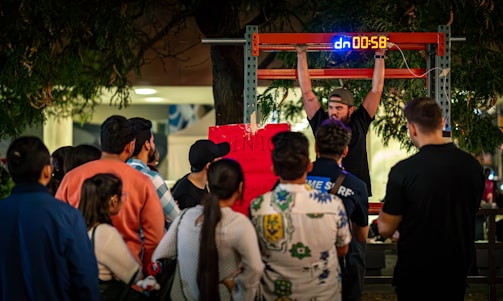 A man is performing a challenge where he hangs from a bar with a digital timer above him. A crowd gathers around, observing with interest and curiosity. The scene takes place outdoors, possibly at a fair or public event, with trees and lights in the background.