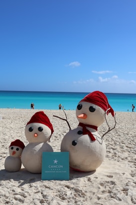 Three sand snowmen with red hats and scarves stand on a sandy beach near the turquoise ocean under a clear blue sky. A sign with 'Cancun Iberostar Selection' is placed in front of them. The scene is bright and sunny, with a few people visible in the background near the water.