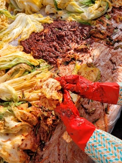 A pair of hands wearing red gloves is preparing kimchi by mixing cabbage with a red chili paste. The image features napa cabbage leaves, some of which are already coated with the spicy mixture, while a large amount of red chili paste is visible on a plastic surface beneath.