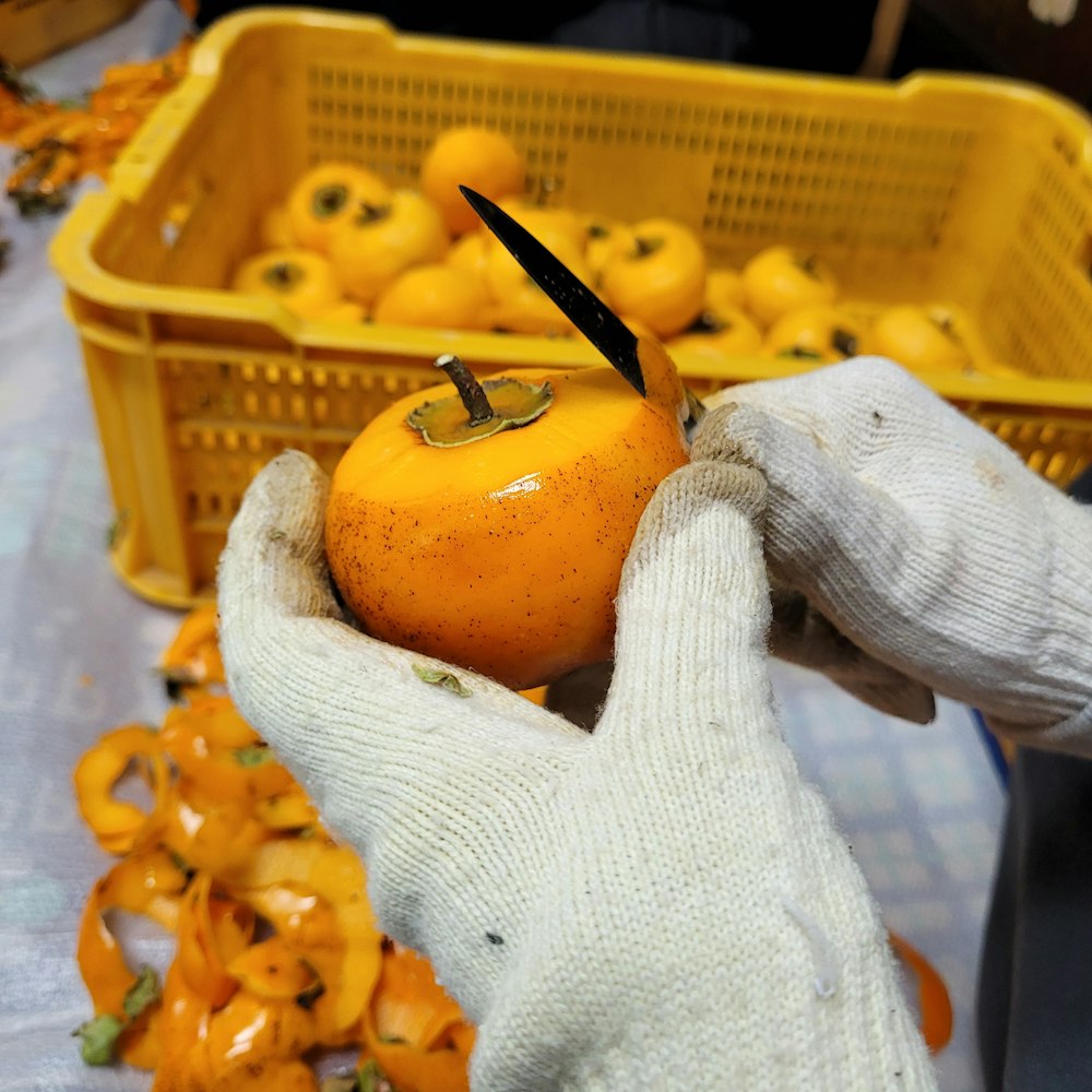 a pair of gloves holding an orange in front of a basket of oranges