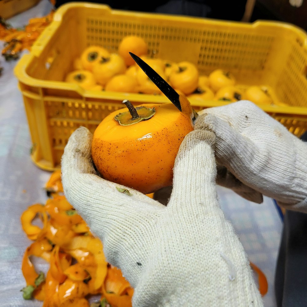 un par de guantes sosteniendo una naranja frente a una canasta de naranjas