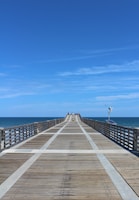 A long wooden pier extends into the ocean under a clear blue sky. Rails line both sides of the pier, and a small number of people are visible at the far end. The ocean is calm and stretches to the horizon. A weather vane equipped with solar panels is mounted on one side of the pier.