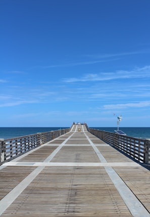 A long wooden pier extends into the ocean under a clear blue sky. Rails line both sides of the pier, and a small number of people are visible at the far end. The ocean is calm and stretches to the horizon. A weather vane equipped with solar panels is mounted on one side of the pier.