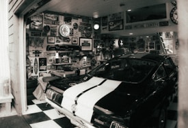 A black car with white racing stripes is parked inside a garage filled with vintage signage, posters, and memorabilia. The walls are adorned with automotive-themed decorations, including old license plates, car emblems, and logos. The checkered flooring adds to the retro atmosphere.