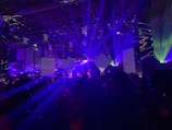 A dynamic scene filled with vibrant laser lights and geometric shapes projected in various colors, creating an energetic and immersive atmosphere. The room is dark, with beams of blue, purple, and green lights cutting through the space, illuminating parts of the audience and stage area.