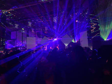 A dynamic scene filled with vibrant laser lights and geometric shapes projected in various colors, creating an energetic and immersive atmosphere. The room is dark, with beams of blue, purple, and green lights cutting through the space, illuminating parts of the audience and stage area.