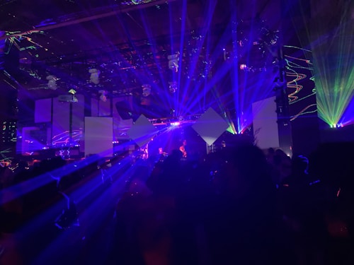A dynamic scene filled with vibrant laser lights and geometric shapes projected in various colors, creating an energetic and immersive atmosphere. The room is dark, with beams of blue, purple, and green lights cutting through the space, illuminating parts of the audience and stage area.