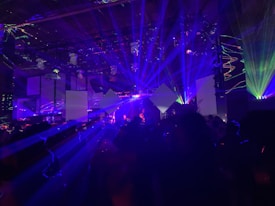 A dynamic scene filled with vibrant laser lights and geometric shapes projected in various colors, creating an energetic and immersive atmosphere. The room is dark, with beams of blue, purple, and green lights cutting through the space, illuminating parts of the audience and stage area.