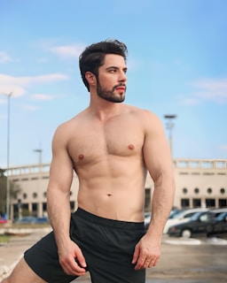 A shirtless man with a well-groomed beard and styled hair stands confidently outdoors. He is wearing black shorts, and a mix of urban and natural elements like parked cars, a building, and a clear blue sky serve as the backdrop.