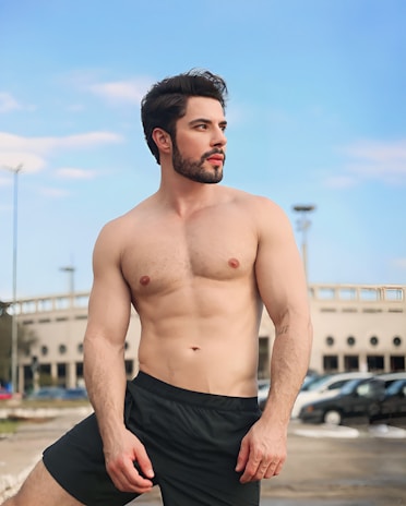 A shirtless man with a well-groomed beard and styled hair stands confidently outdoors. He is wearing black shorts, and a mix of urban and natural elements like parked cars, a building, and a clear blue sky serve as the backdrop.