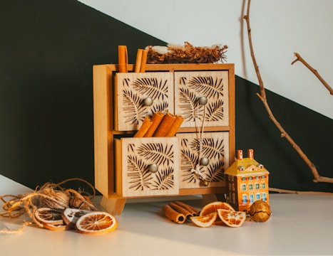 A small wooden cabinet featuring intricately carved drawer fronts is displayed. Cinnamon sticks protrude from the drawers, adding a rustic touch. Nearby, dried orange slices, a decorative miniature house, and a natural branch create an autumnal or holiday atmosphere.