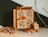 A small wooden cabinet featuring intricately carved drawer fronts is displayed. Cinnamon sticks protrude from the drawers, adding a rustic touch. Nearby, dried orange slices, a decorative miniature house, and a natural branch create an autumnal or holiday atmosphere.