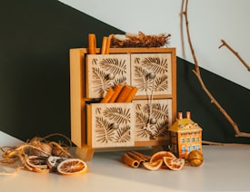 A small wooden cabinet featuring intricately carved drawer fronts is displayed. Cinnamon sticks protrude from the drawers, adding a rustic touch. Nearby, dried orange slices, a decorative miniature house, and a natural branch create an autumnal or holiday atmosphere.