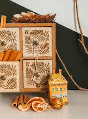 A decorative set-up featuring a wooden storage box with intricate leaf patterns on its drawers, accompanied by a small, colorful ceramic house. Cinnamon sticks, dried orange slices, and possibly nuts are arranged in front, with a natural twig and a small wreath on top of the box adding an earthy touch.