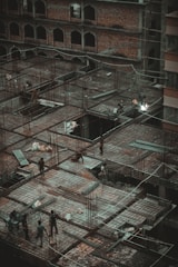 A construction site with workers actively engaged in building a structure. The site consists of metal frameworks and unfinished concrete slabs laid out in a grid-like pattern. Several construction materials, including metal beams and rebar, are scattered across the site. The background features a brick building with multiple arches and open windows.