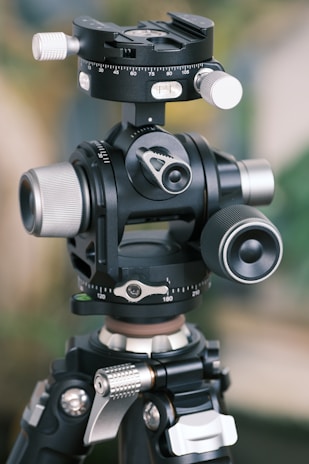 A detailed close-up view of a sophisticated tripod head with various adjustment knobs and dials. The design shows precision engineering, featuring multiple levels and a rotating mechanism for angular adjustments.