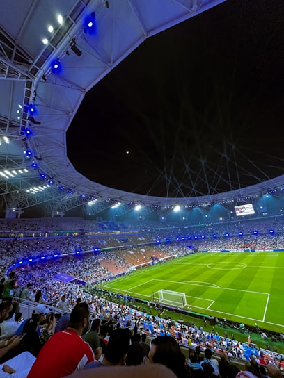 A large, illuminated stadium filled with spectators, with a focus on a well-maintained football field and energetic crowd. Bright stadium lights cast a glow over the scene, enhancing the lively atmosphere as people watch an ongoing sporting event.