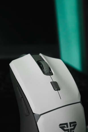 A sleek, modern computer mouse predominantly white with gray accents. It features a scroll wheel and several buttons, indicating a design suitable for gaming or productivity. The mouse is positioned against a dark background with a teal vertical light strip.