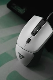 A sleek white gaming mouse with a modern design rests on a subtle green and grey patterned surface. The mouse features a scroll wheel and a logo on its surface.