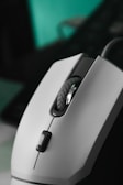 A close-up view of a computer mouse with a sleek design. The mouse is predominantly white with a textured scroll wheel and a dark clickable button. The background is blurred, highlighting the mouse as the focal point.