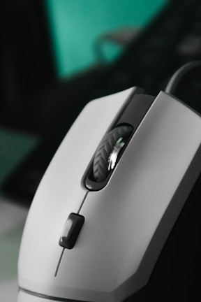 A close-up view of a computer mouse with a sleek design. The mouse is predominantly white with a textured scroll wheel and a dark clickable button. The background is blurred, highlighting the mouse as the focal point.