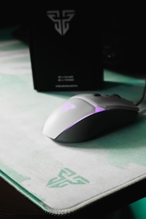 A sleek, white gaming mouse with purple LED lighting sits on a light green and white mouse pad, both featuring a logo. In the background, a black box with a similar logo is visible.