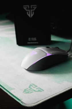 A sleek, white gaming mouse with purple LED lighting sits on a light green and white mouse pad, both featuring a logo. In the background, a black box with a similar logo is visible.