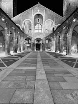 A black-and-white photograph of a historic, symmetrical architectural structure featuring arches and columns. The perspective creates a converging line, drawing the eye towards the building at the center. The paved courtyard is accentuated by geometric patterns, and the structure is illuminated dramatically against a dark night sky.