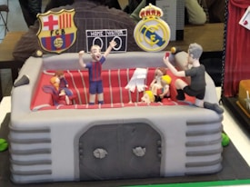 A cake designed to look like a mini football stadium with animated figures, featuring one player in a blue and red striped jersey celebrating with raised arms. Other figures are seated or standing nearby. There are logos resembling those of famous football clubs on either side. The seating area of the cake is detailed with red seats and figures dressed in different colors.