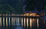 A series of charming wooden bungalows rests on a pier extending over a calm body of water. The scene is set against a backdrop of lush, forested cliffs. Warm lights illuminate the bungalows, casting long, colorful reflections onto the tranquil water's surface.